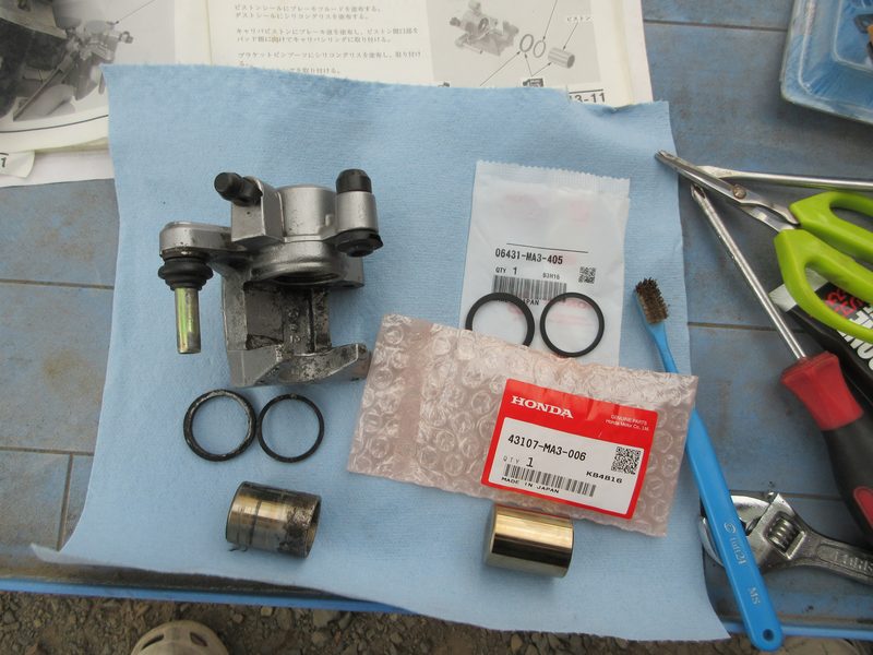 ホンダ・ジョーカー90のキャリパー本体、取り外したピストンシール類と新品のホンダ純正ピストンとピストンシールキット